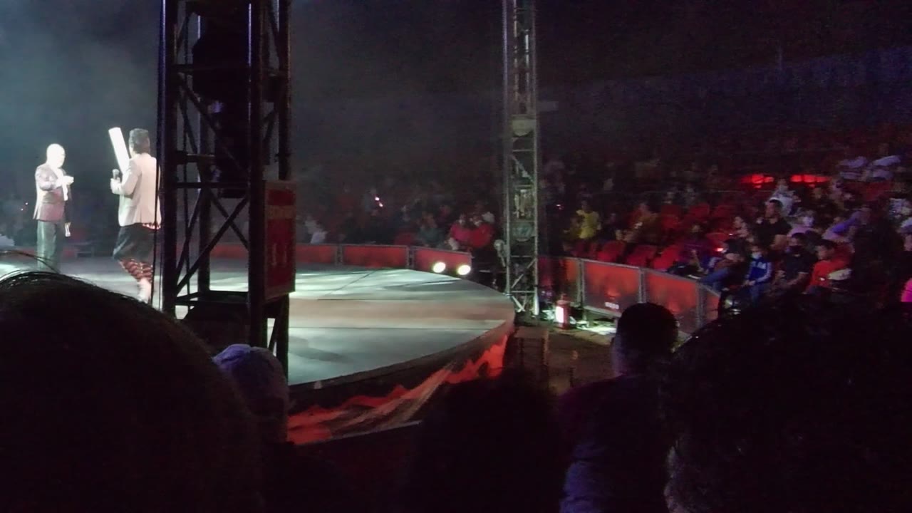 Two performers on a stage in San Jose Del Cabo, Baja California Sur, with one holding a microphone, another holding a white prop, red audience seats, and foreground spectator silhouettes.