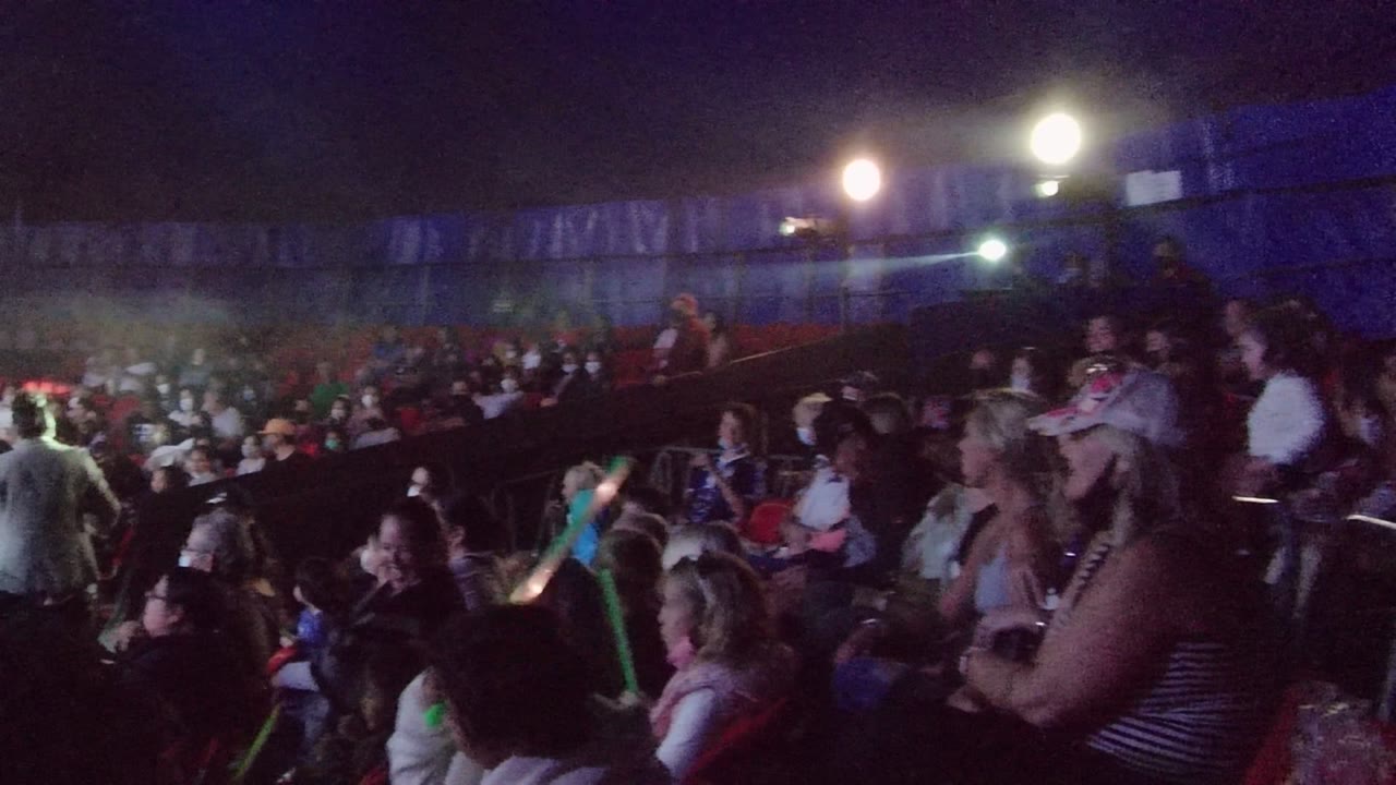 Audience members sit in red chairs in a dim San Jose Del Cabo theater while a lit stage area appears at the left edge.