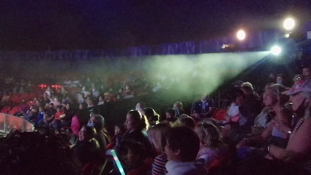 A San Jose Del Cabo nighttime concert audience sits in red stadium seating while a green spotlight beam and haze spread above the crowd.