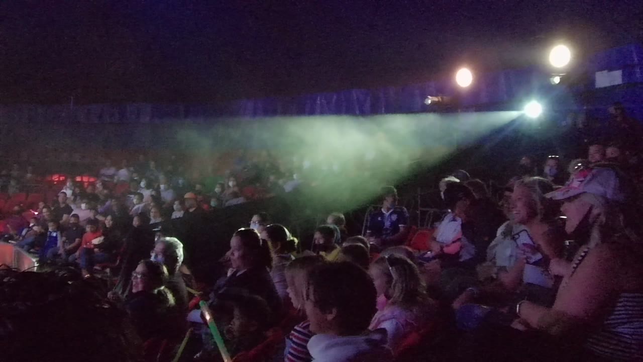 Seated spectators watch a nighttime performance with smoke and blue lighting.