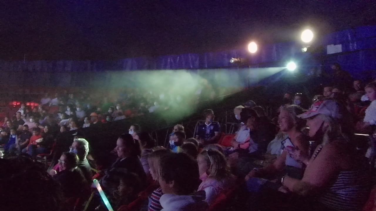 A child and adults sit in the foreground while the San Jose Del Cabo concert crowd extends into the background under green haze and blue light.