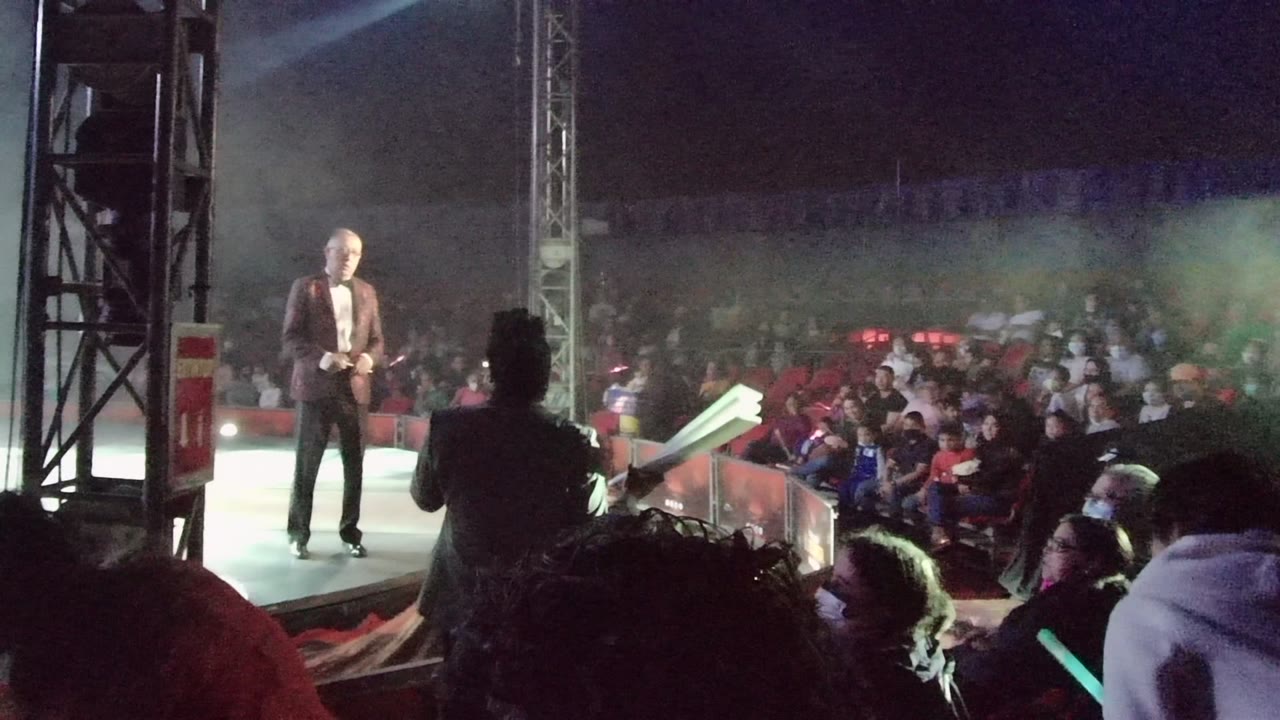 A suited performer stands on a lit stage in San Jose del Cabo while a seated audience watches from the darkened venue.