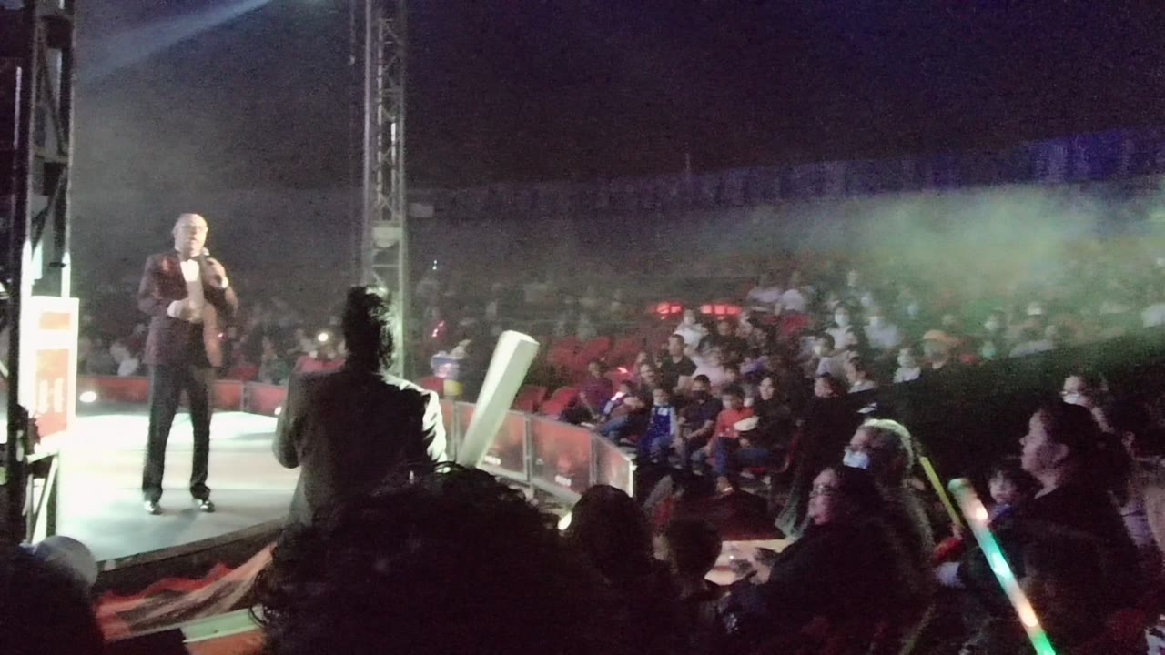 The performer holds a microphone on the San Jose del Cabo stage as spectators sit in rows and glow sticks appear near the foreground.