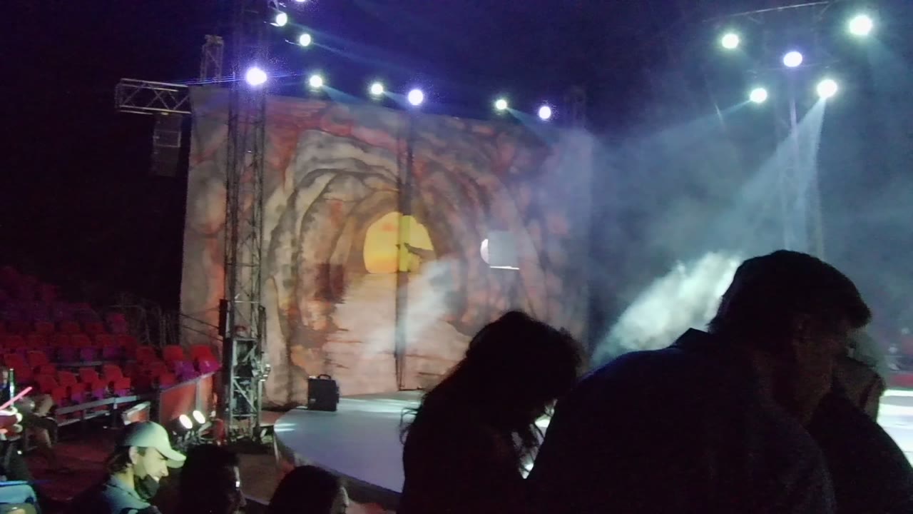 Audience silhouettes in San Jose del Cabo face a smoky stage with blue-white lights, red seats at left, and a cave-themed backdrop with a yellow circle.