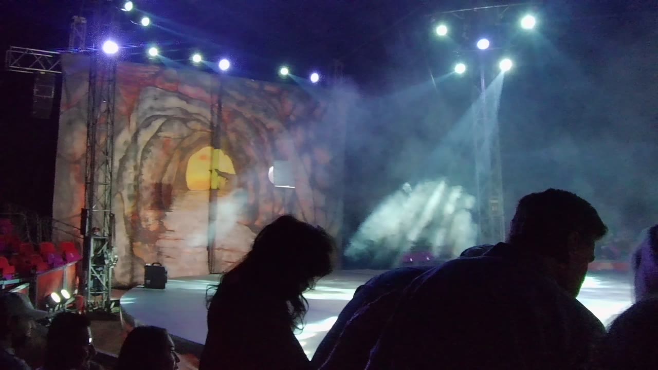 Dark audience figures sit before a lit stage with smoke and a painted cave backdrop.