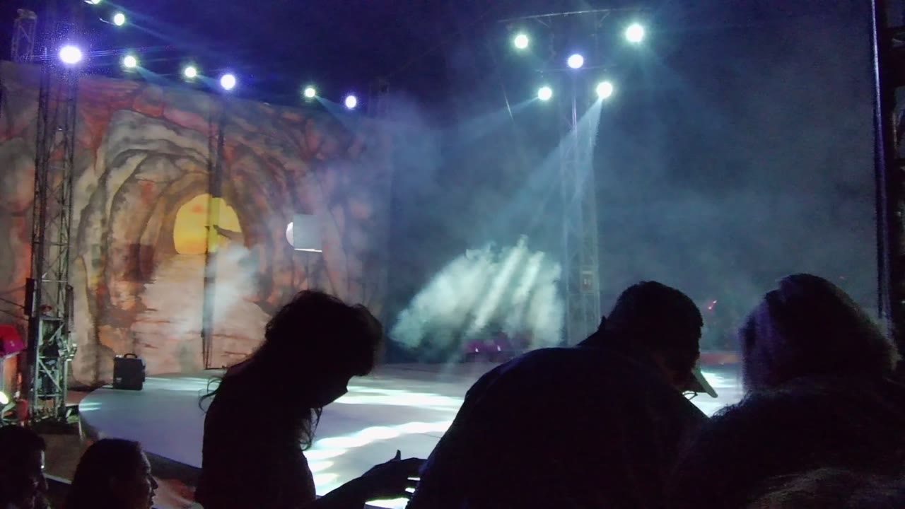 Audience silhouettes lean forward in San Jose del Cabo while bright beams cut through smoke over the open stage and painted backdrop.