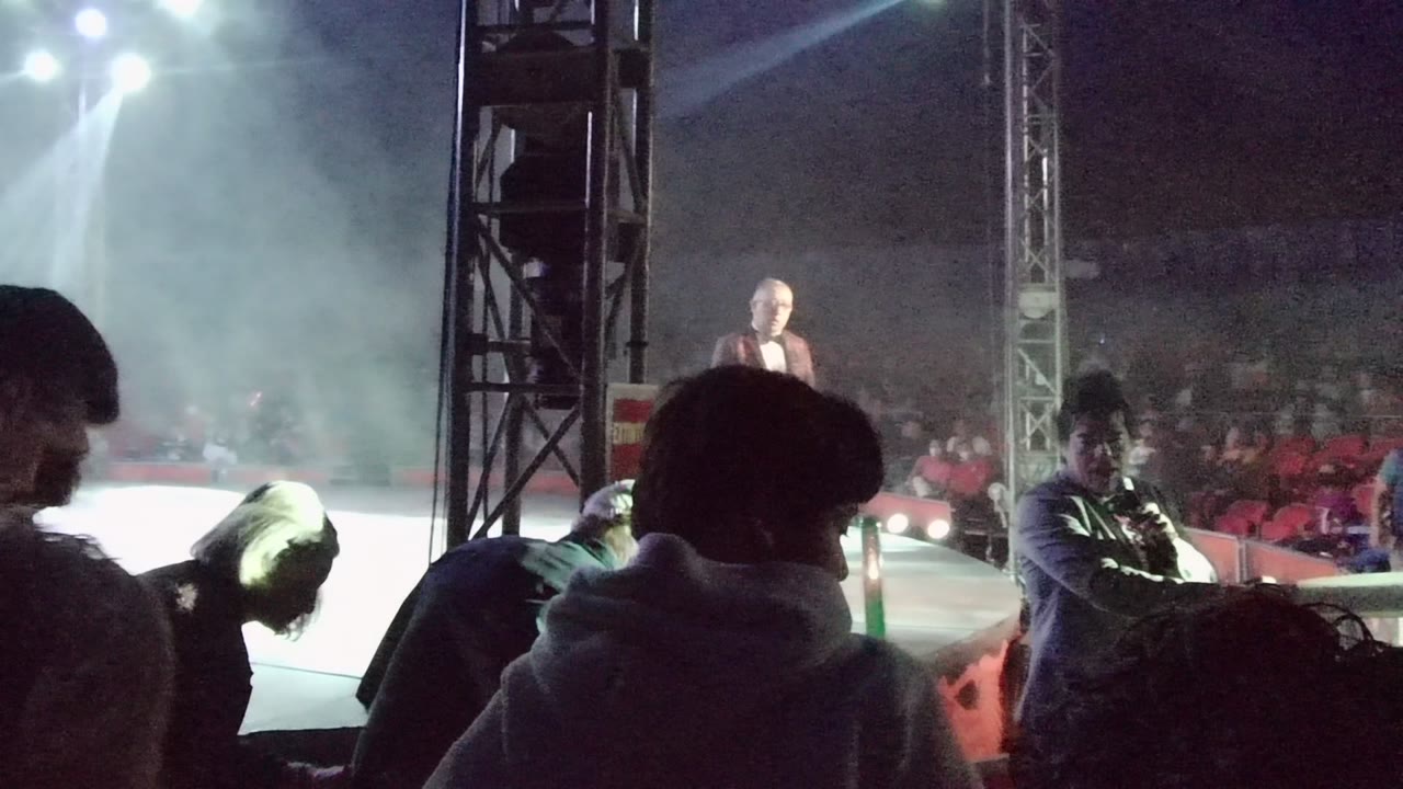A camera operator or crew member appears near the stage edge as the suited performer stands farther back under smoke and bright lights in San Jose del Cabo.