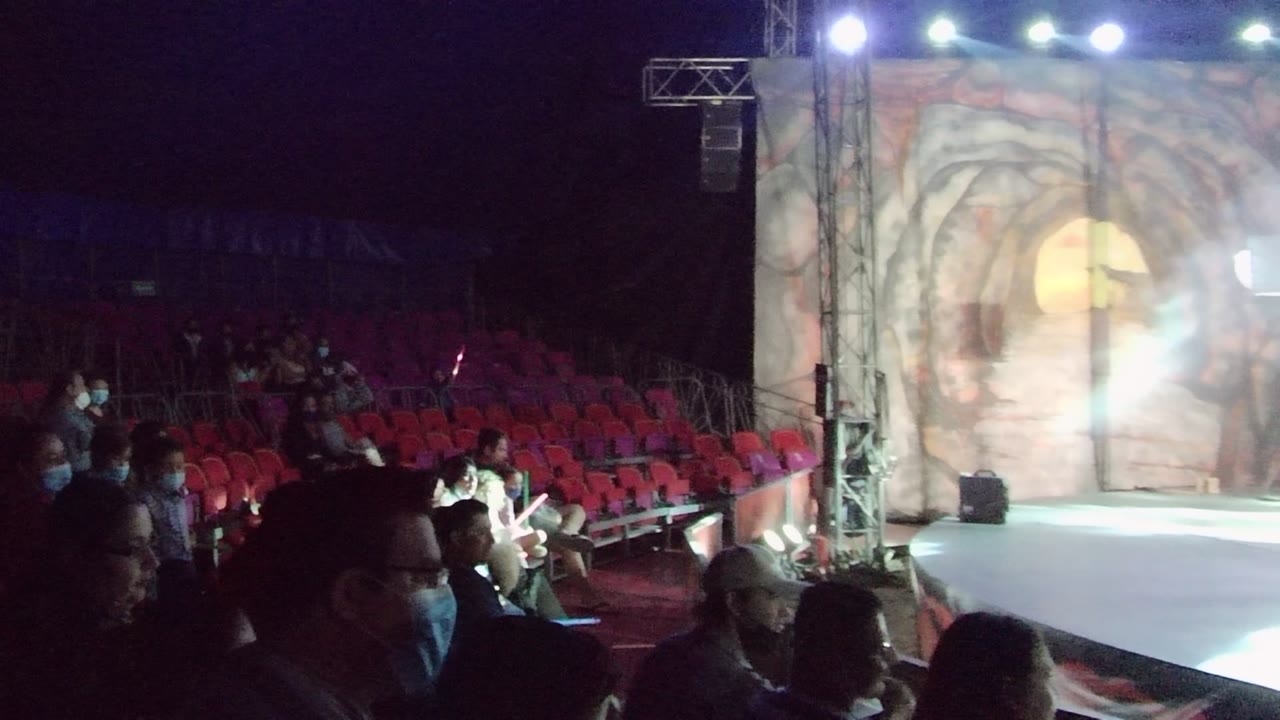 At 0:00 in San Jose del Cabo, audience members sit in red seats while a bright white stage and cave-themed painted backdrop fill the right side of the frame.