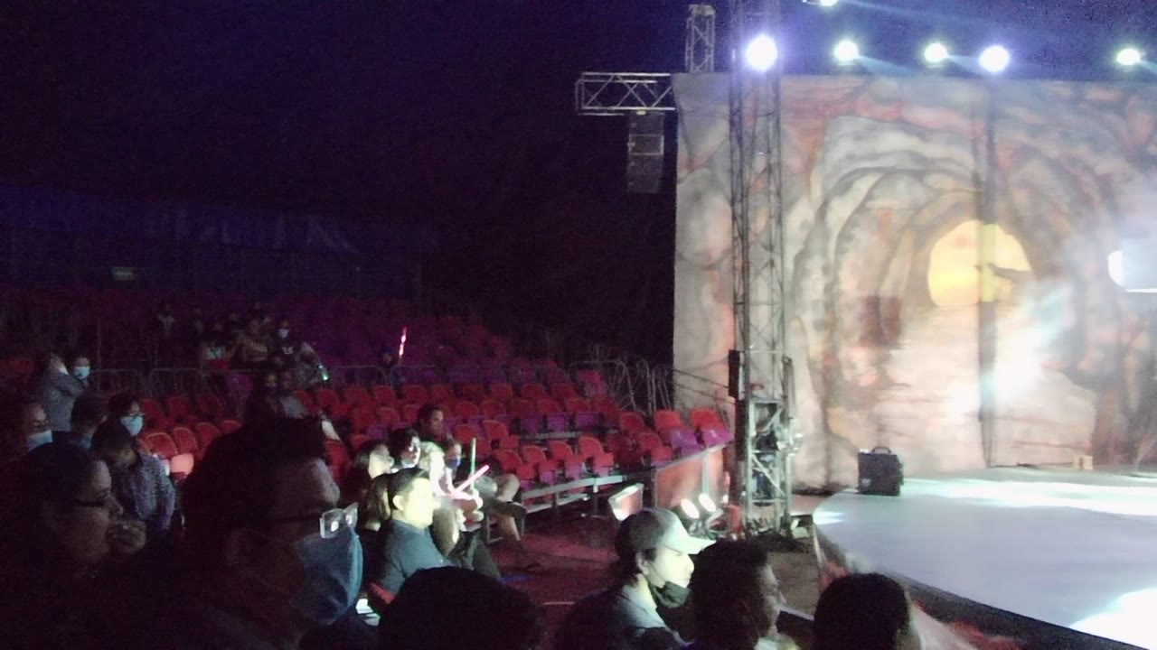 Red theater seating and spectators face a bright stage with smoke near the backdrop.