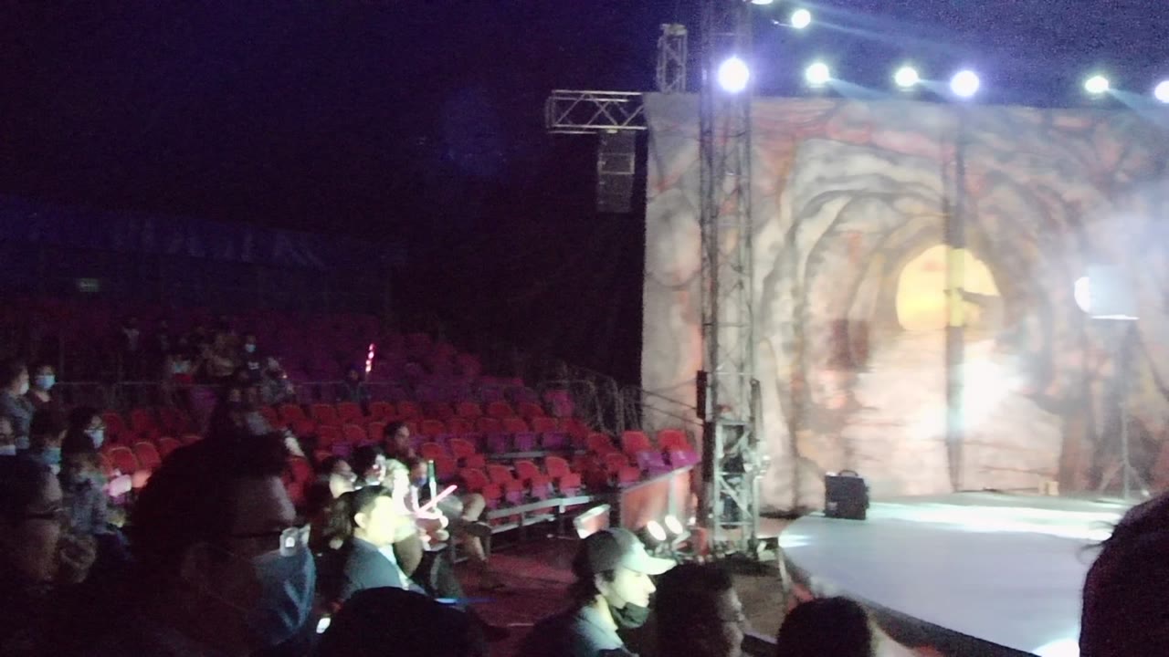 At 0:02 in San Jose del Cabo, dark foreground silhouettes frame the audience rows while the illuminated stage and cave-themed backdrop remain the main background elements.