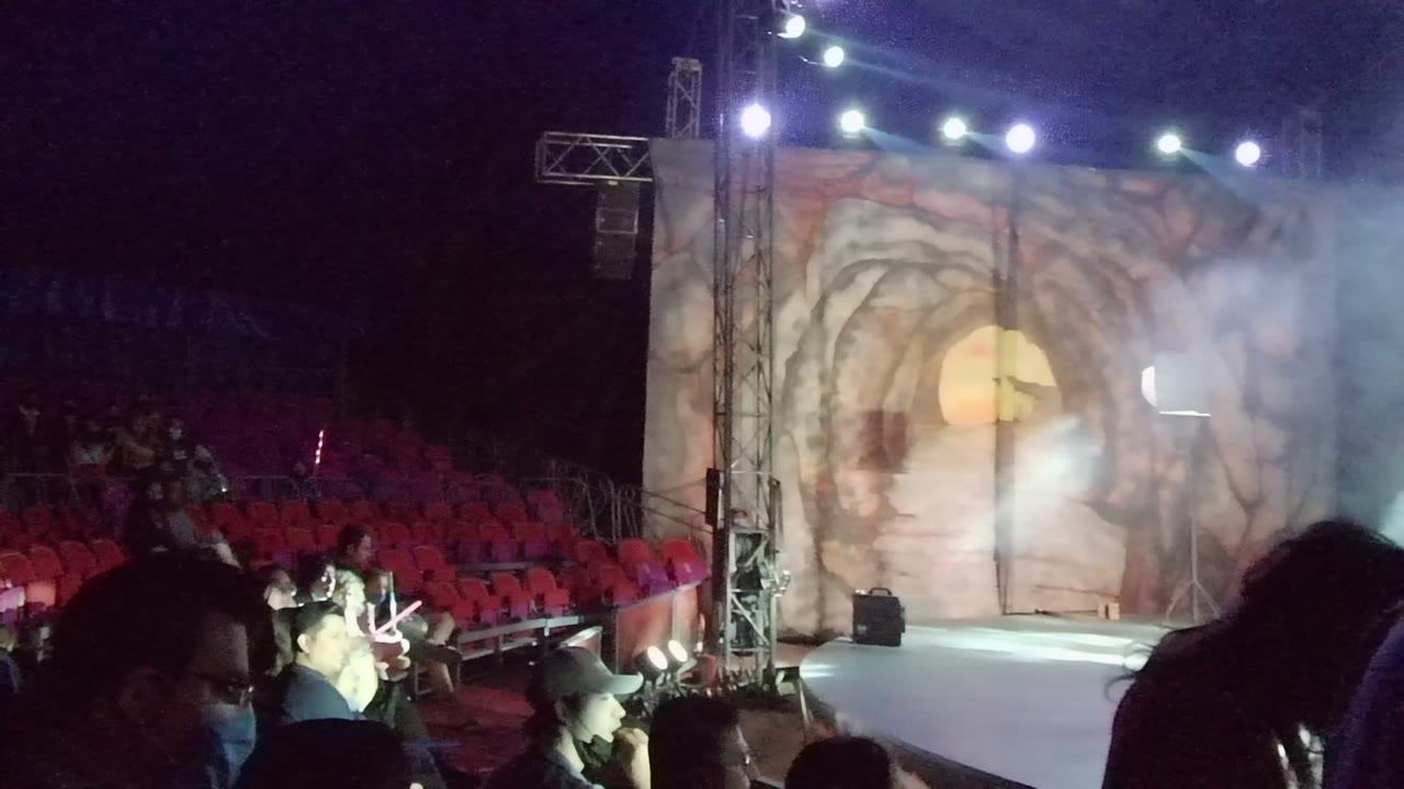 Smoke drifts across a stage while audience members sit in red chairs.