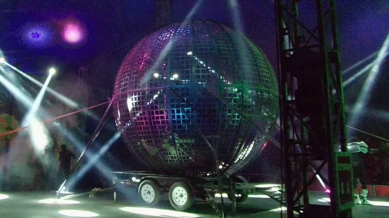 A giant grid-pattern sphere on a wheeled trailer is lit by magenta, green, and white stage beams in San Jose Del Cabo, with smoke and small people visible near the stage edges.