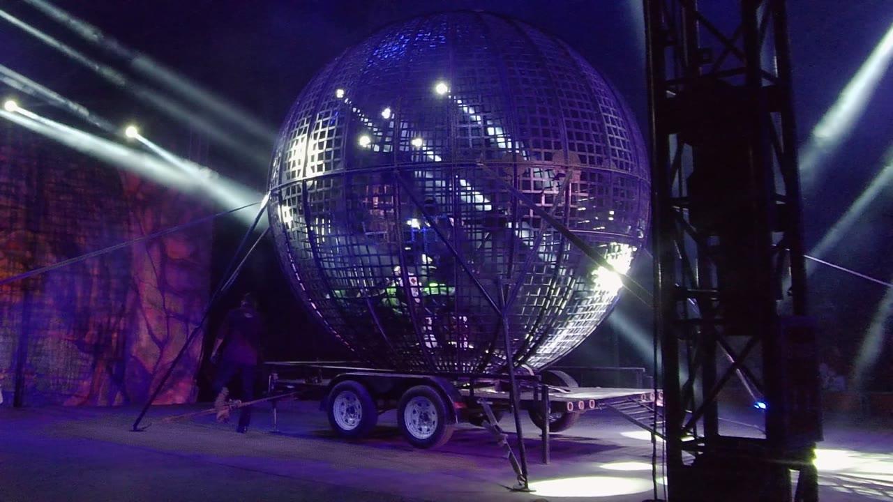 A giant illuminated grid-pattern sphere on a trailer in San Jose Del Cabo, Mexico, with purple-blue stage lighting, white beams, truss framing, and no visible people.