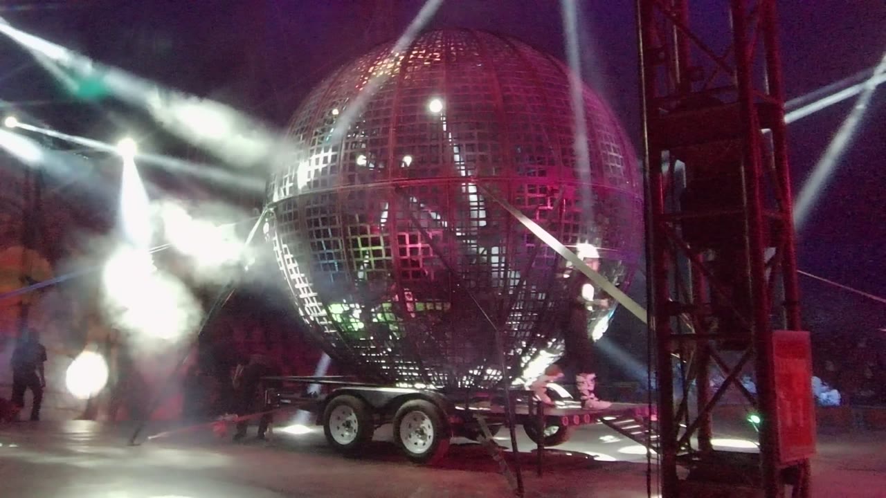 A burst of smoke and bright white stage lights flare beside the red-lit grid sphere on its trailer in San Jose Del Cabo, Mexico, with people faintly visible near the stage.
