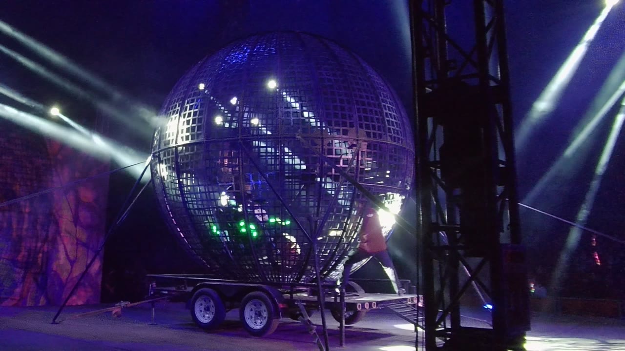 Large grid-patterned reflective sphere on a trailer in San Jose Del Cabo, Mexico, with white beams, blue-purple lighting, and a person partly visible near the right side.