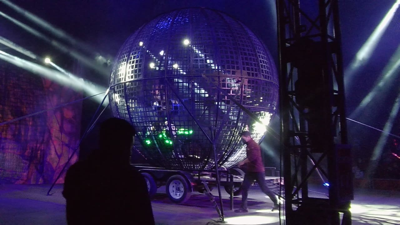 The reflective grid sphere sits on its trailer in San Jose Del Cabo, Mexico, as a person moves at the right edge and a dark silhouette crosses the lower foreground.