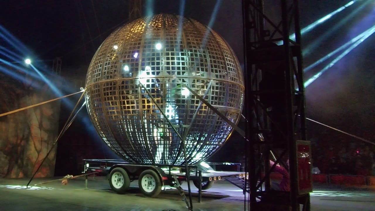 A large golden metal grid sphere on a trailer is lit by blue and white spotlights at a nighttime event in San Jose del Cabo, with no visible people in the foreground.