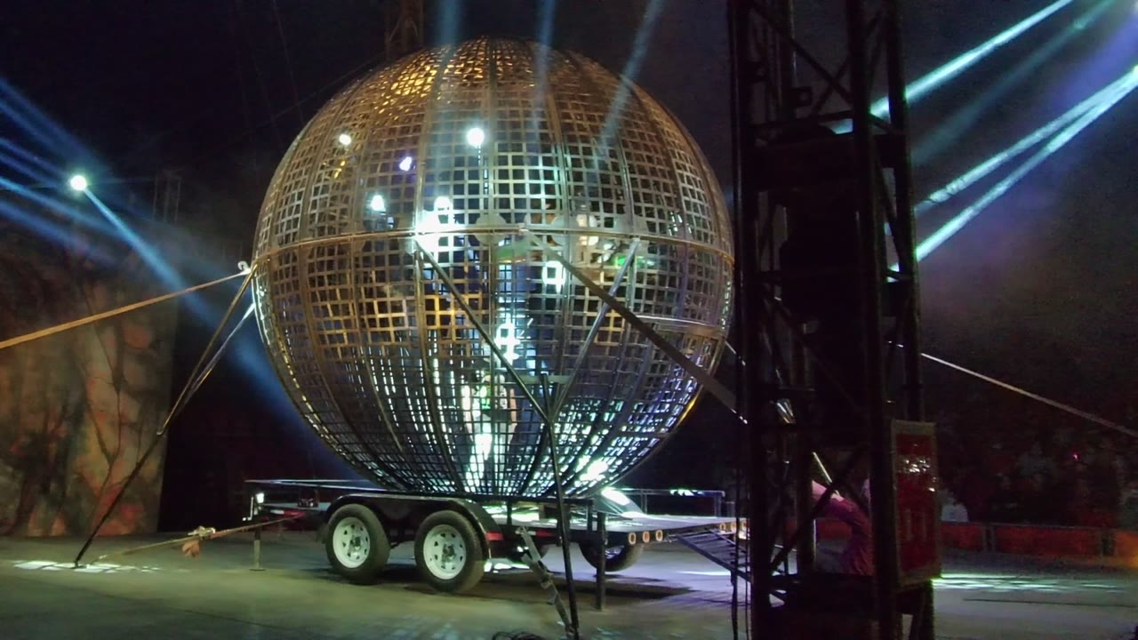 A giant metal sphere on a wheeled trailer is framed against crossing blue light beams and a dark stage backdrop during the San Jose del Cabo nighttime event.