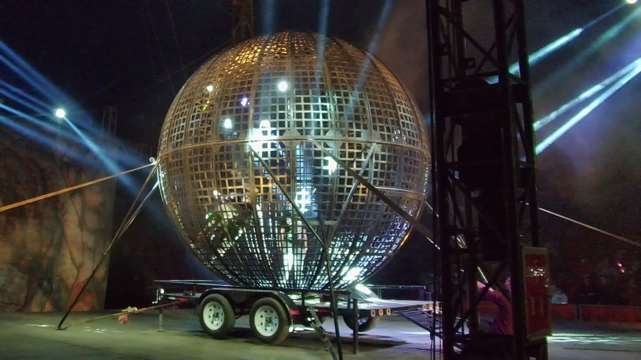 A giant golden open-grid sphere on a trailer is lit by blue-white stage beams in San Jose del Cabo, Mexico, with smoke behind it and no visible people.