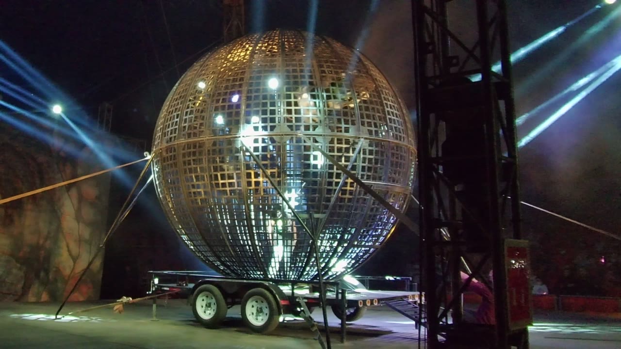 Large golden sphere on a trailer with smoke and diagonal stage lights.