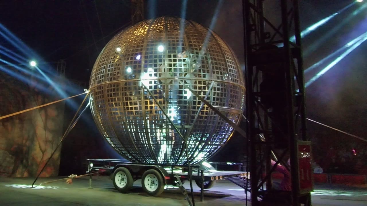 A lit golden spherical prop on a trailer fills the frame in San Jose del Cabo, Mexico, with a black stage truss at the right edge, bright reflections on the lattice, and no visible people.