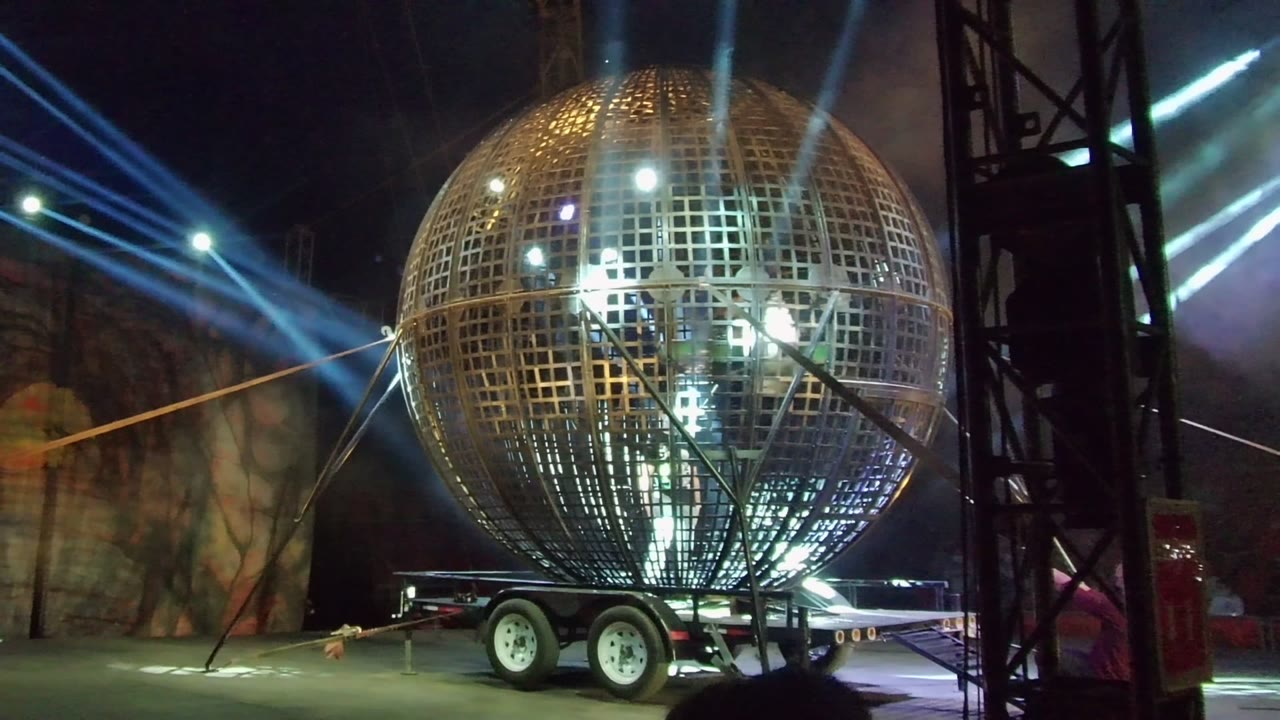 A golden metal-grid sphere on a trailer is lit by blue-white spotlights during a nighttime event in San Jose del Cabo, with people visible inside and a dark truss at the right edge.