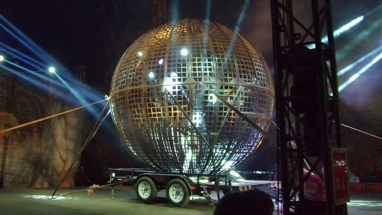 Illuminated globe-like stage structure with people inside and beams overhead.