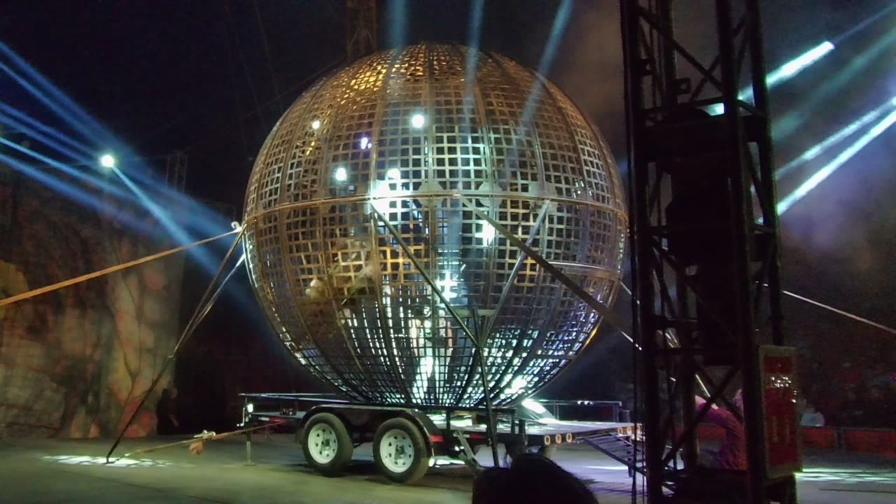 A trailer-mounted golden sphere remains centered in the nighttime San Jose del Cabo performance area, with diagonal light beams behind it and human figures visible inside the grid.