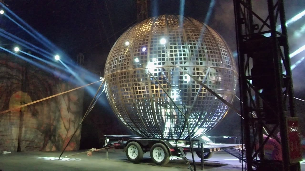 A large metallic grid sphere on a wheeled trailer is lit by blue-white stage beams in San Jose Del Cabo, with black truss at the right edge and no visible people.
