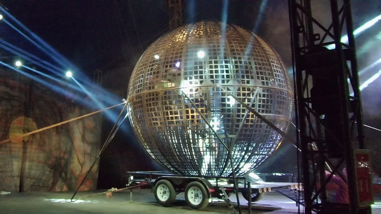 A large mirrored grid sphere sits on the trailer in San Jose Del Cabo, with diagonal support lines crossing the frame, bright lights inside the sphere reflections, and no visible people.