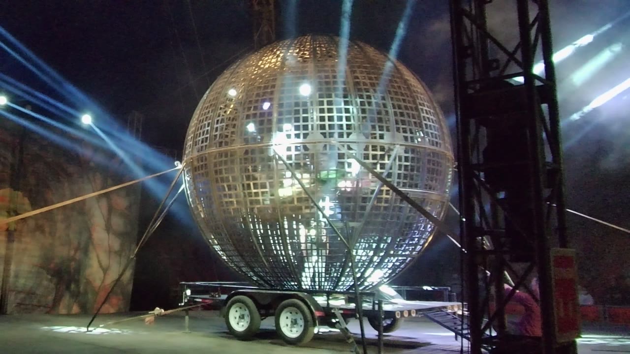 Large spherical metal grid structure on a trailer in San Jose del Cabo at night, with blue-white stage beams behind it and no visible people.