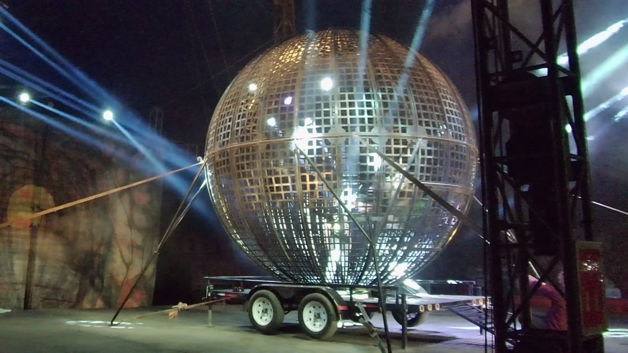 Large illuminated sphere on a wheeled trailer in San Jose del Cabo, framed with dark vertical truss at right, crossing support lines, blue beams, and no visible people.