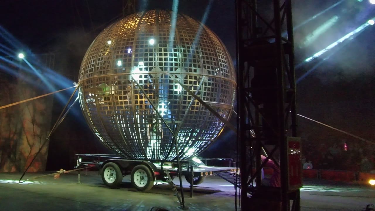 Large golden lattice sphere on a trailer in San Jose Del Cabo, Baja California Sur, with blue beams, haze, stage truss, and no visible people.