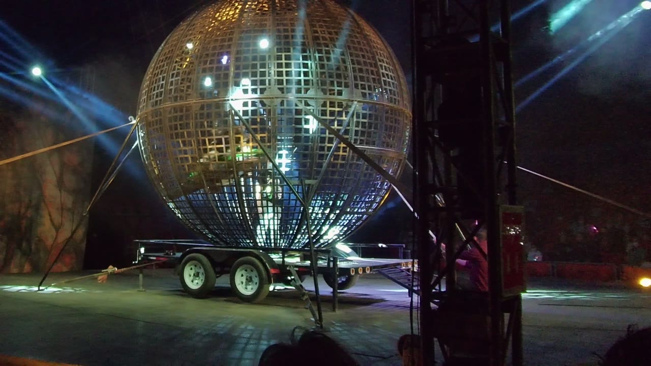 A large metal grid sphere on a trailer is lit from inside at night in San Jose del Cabo, with stage beams and haze behind it.