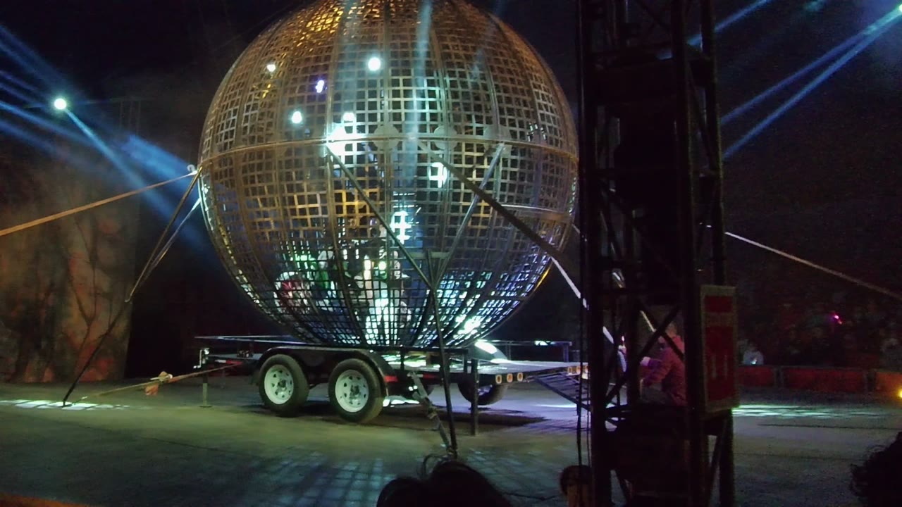 Large metallic grid sphere on a trailer in San Jose del Cabo at night, with blue-white spotlights crossing behind it and people visible inside the structure.