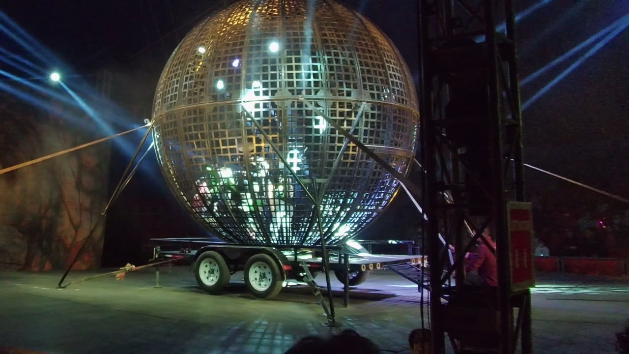 Illuminated metal grid sphere on a wheeled trailer in San Jose del Cabo at night, with blue spotlights crossing the dark background and partial silhouettes near the lower edge.