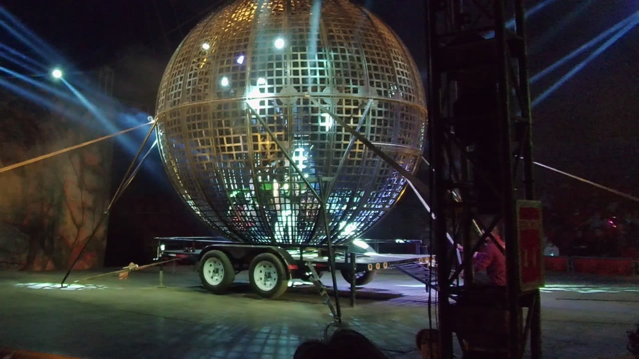 The lit spherical structure in San Jose del Cabo fills the right side of the frame, with an amber beam and blue spotlights visible across the dark outdoor stage background.