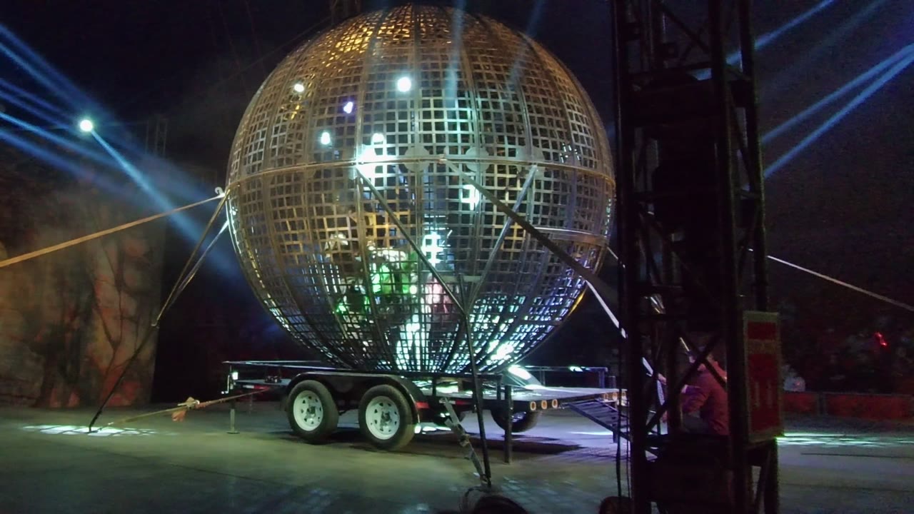 A giant golden grid sphere on a wheeled trailer is lit at night in San Jose del Cabo, with blue spotlights crossing behind it and small people visible inside the frame.