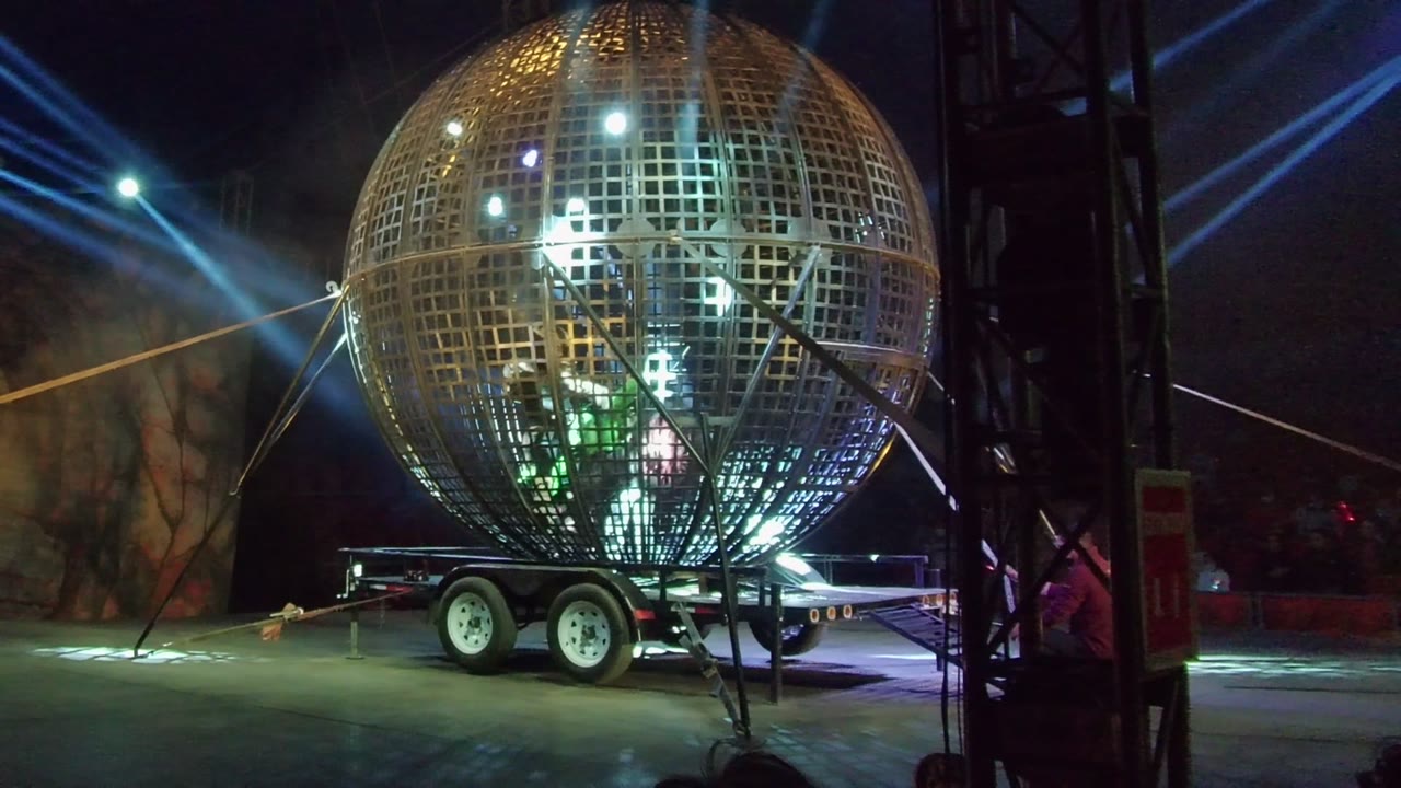 Illuminated metal sphere with people inside and blue beams behind it.