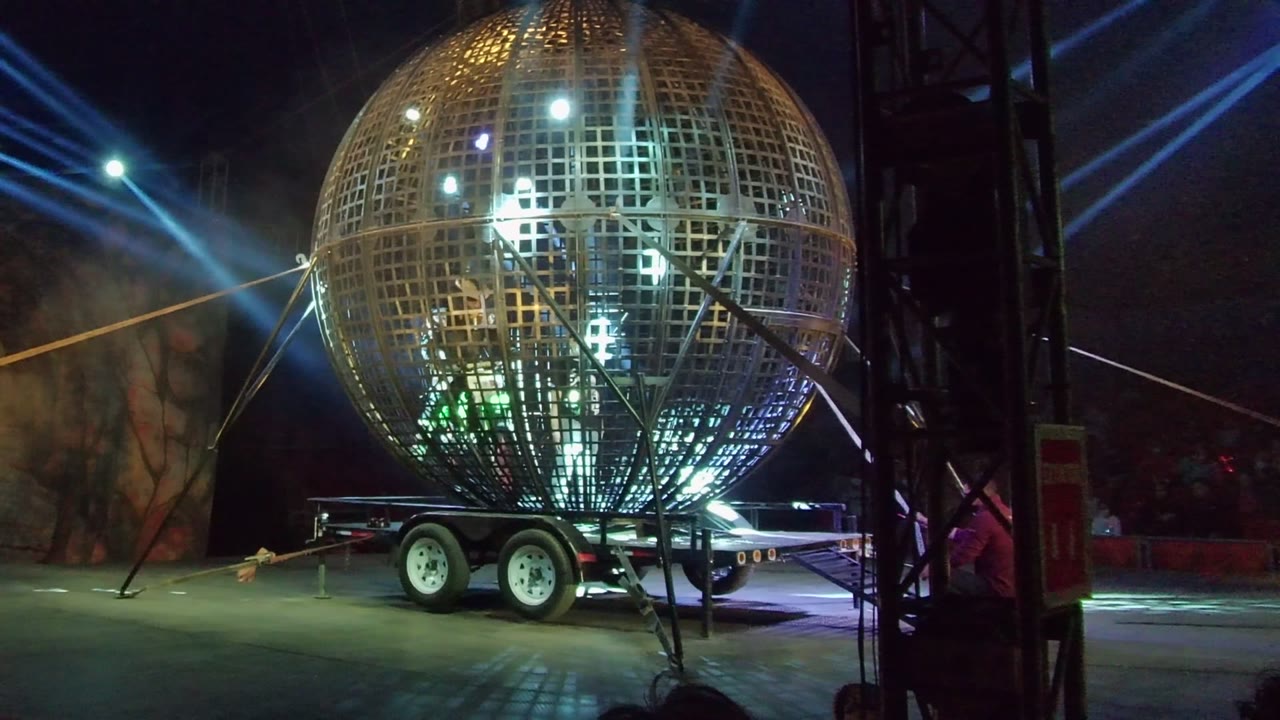 A golden lattice globe on a trailer fills the frame in San Jose del Cabo, with a dark stage truss along the right side and blue light beams angled across the night background.