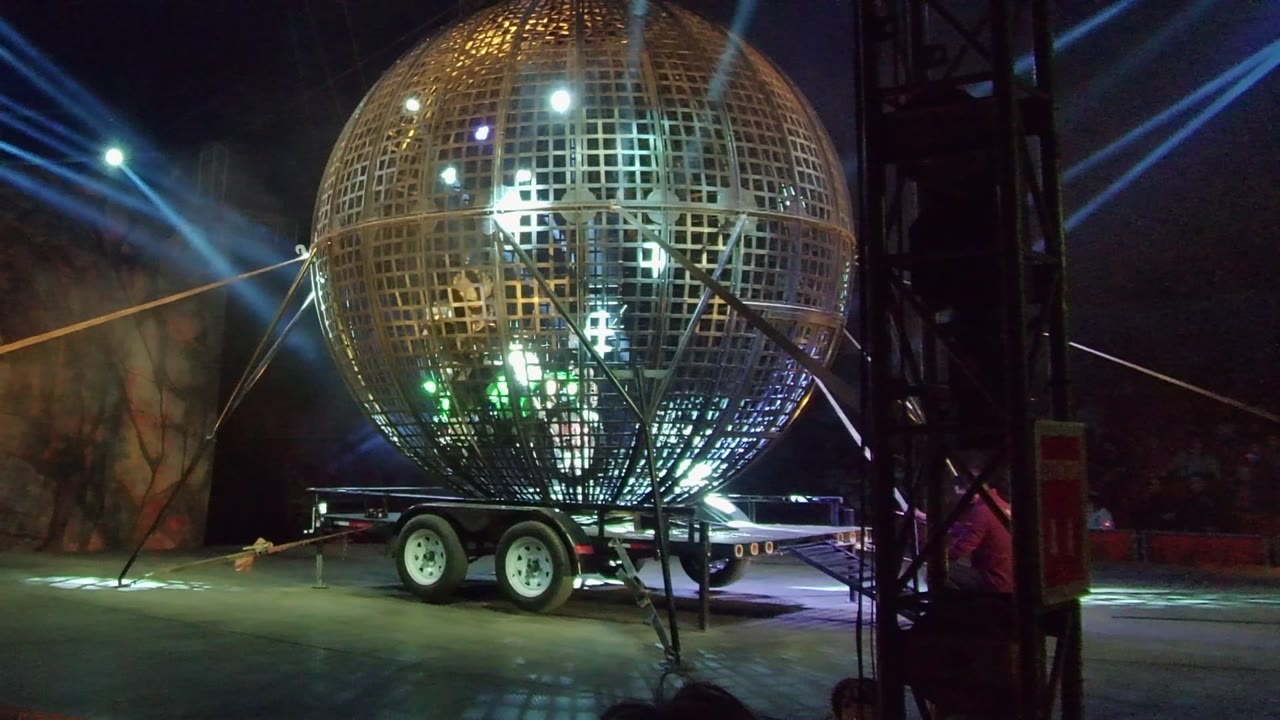 Large metal grid sphere on a wheeled trailer in San Jose del Cabo, with blue spotlights crossing the dark background and no visible people.