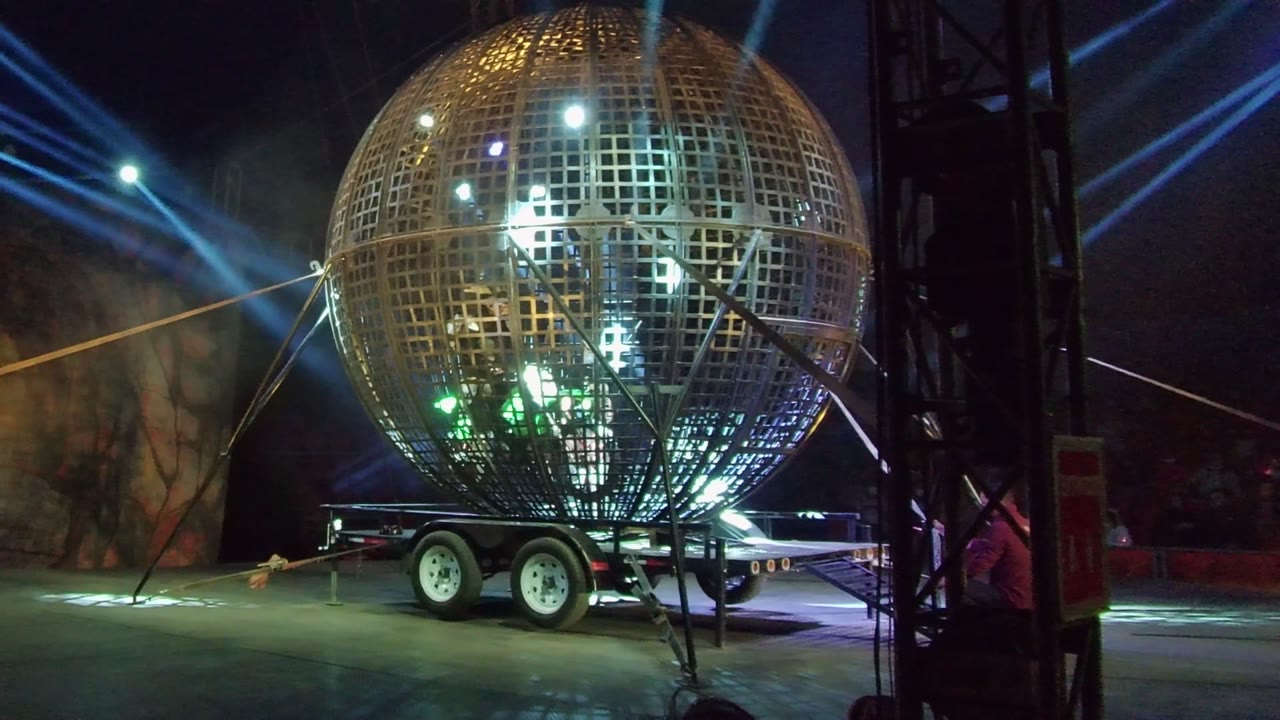 Large grid-pattern sphere on a trailer in San Jose del Cabo, backed by diagonal blue stage beams and dark surroundings, with no visible people.