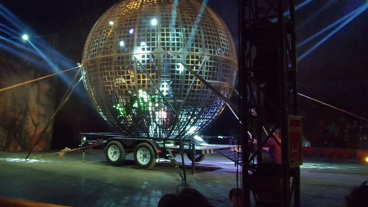 A giant metallic grid sphere mounted on a trailer sits under blue and white stage beams in San Jose Del Cabo, with rigging at the right edge and people faintly visible near the apparatus.