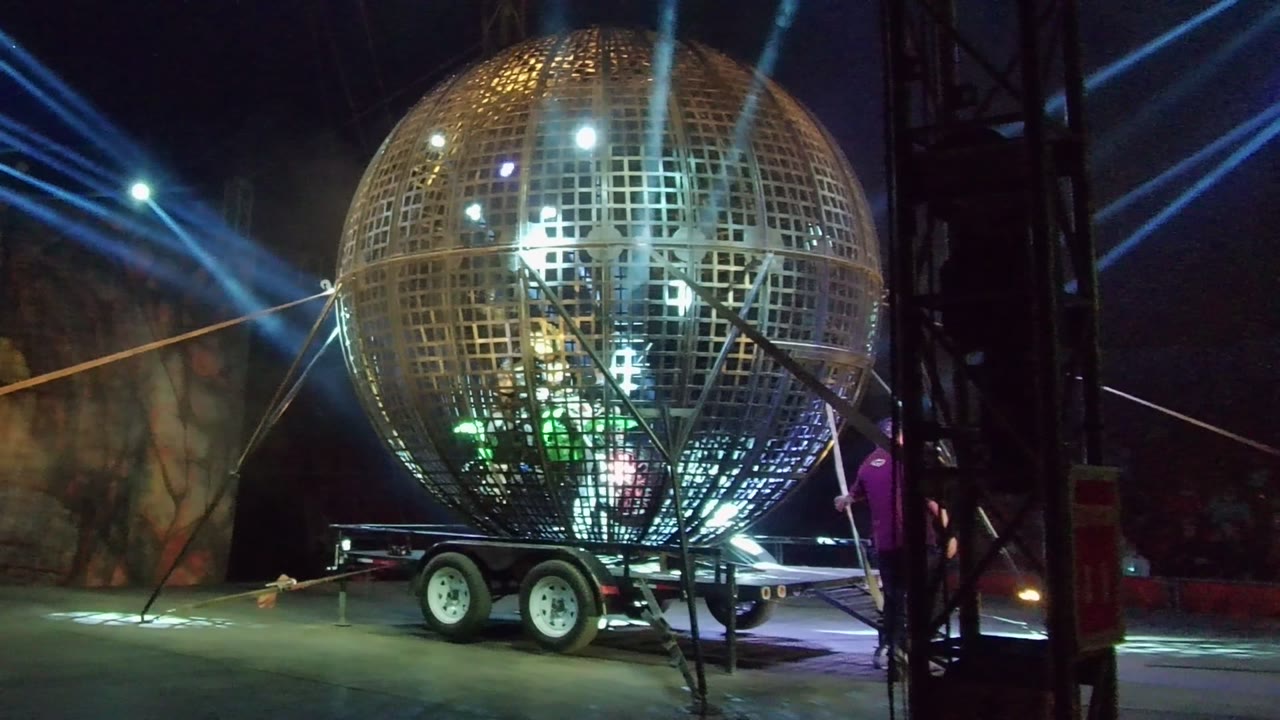 A large grid-patterned spherical structure on a trailer is lit by blue and white beams in San Jose Del Cabo, with stage truss at the right edge and people faintly visible around the structure.