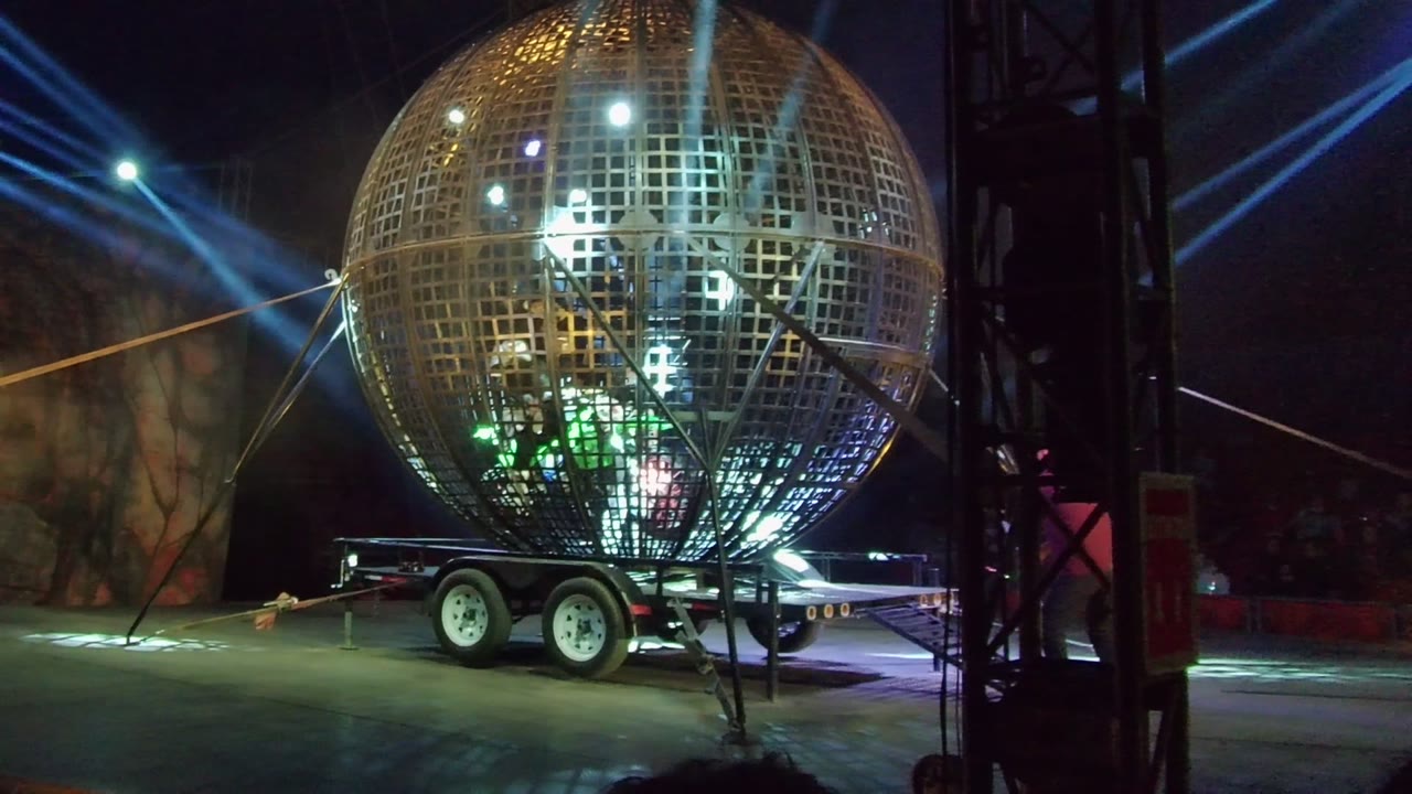 In San Jose Del Cabo, the metal sphere on the trailer shows brighter white points near the top and green light near the lower grid, with diagonal blue beams sweeping behind it and people visible within the lit structure.