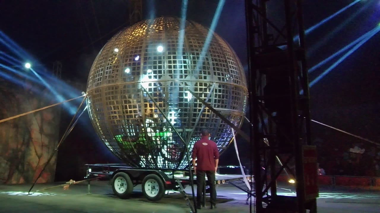 At 0:00 in San Jose del Cabo, a large illuminated mirror ball on a trailer fills the stage area while a person in a red shirt stands with their back to camera beneath blue light beams.
