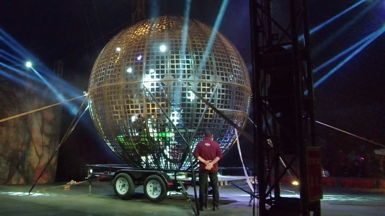 Person stands near a reflective mirror ball trailer as blue beams cross the dark stage.