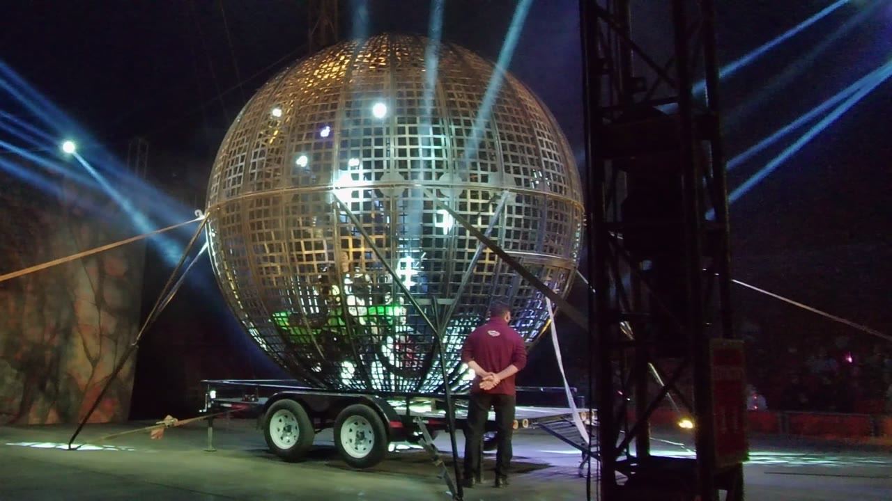 At 0:02, the mirror ball remains centered on its trailer in San Jose del Cabo while the person in red stands at the right edge, partly framed by dark rigging and blue overhead light beams.