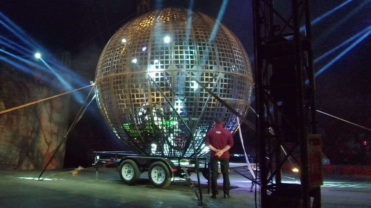 A giant golden spherical stage structure on a trailer stands under blue beams in San Jose del Cabo, with a person in a red shirt near the right side.