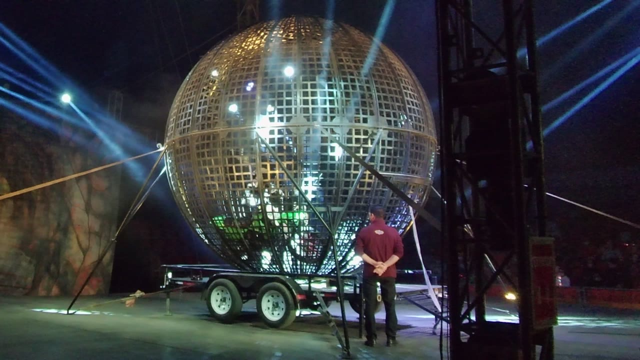 Large golden sphere on stage with blue light beams and a person beside the trailer.